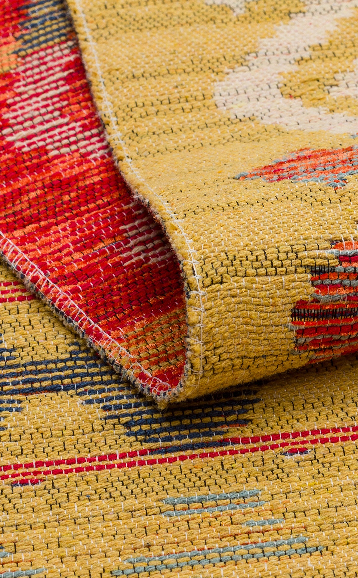 A close-up of the Crimson Dot - Kilim - NMD03 rug shows its folded corner, emphasizing the yellow background with red, blue, and beige patterns as well as its detailed texture and stitching.