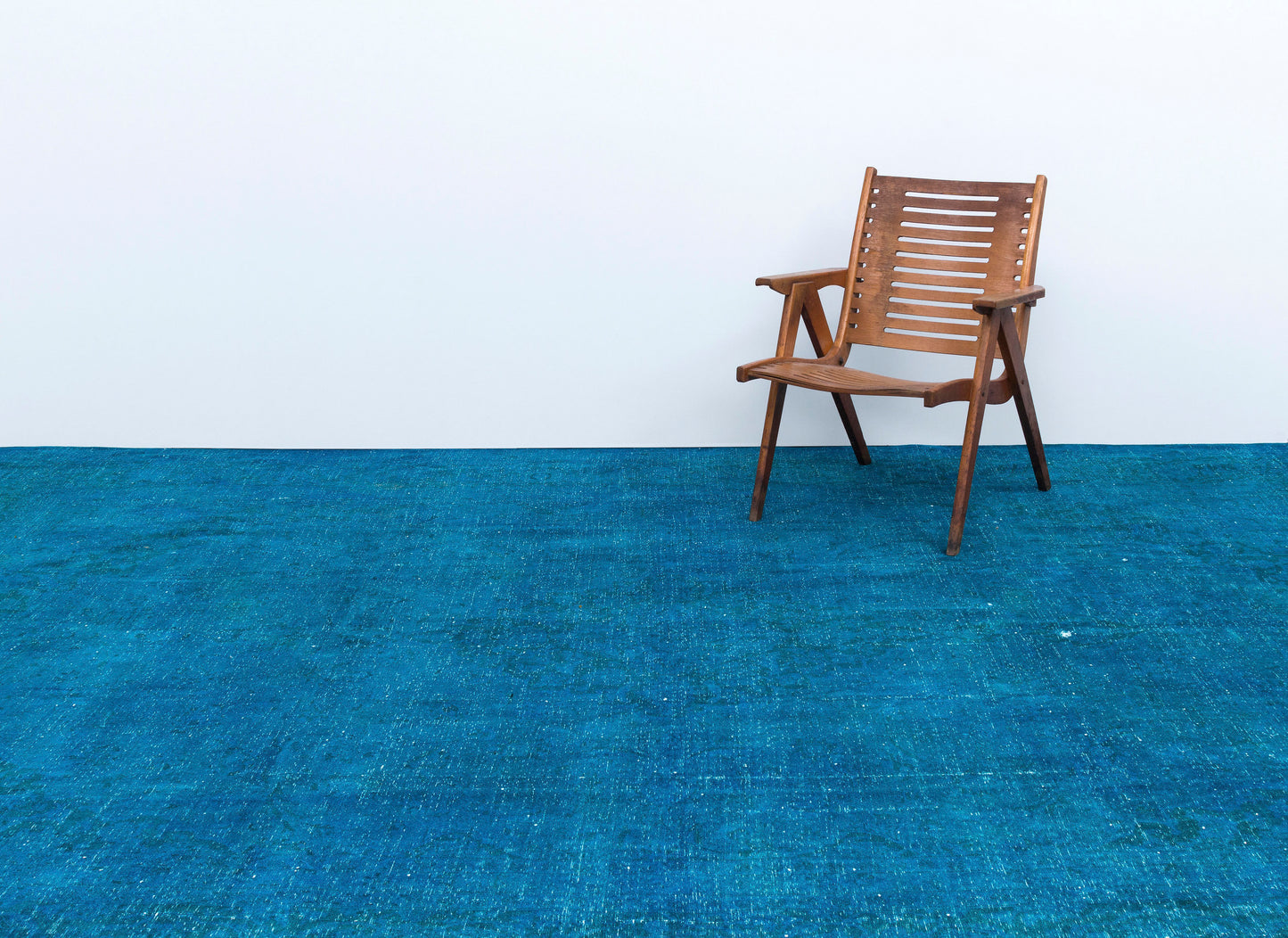 Solid blue distressed-texture rug with a wooden slatted chair, set against a white wall.