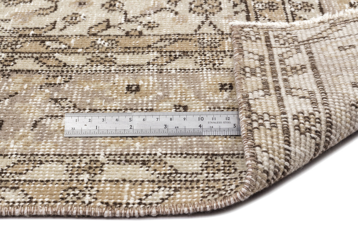 A metal ruler sits on the Coaluon - Vintage Floral Rug (5'8 x 9'1), its corner lifted to display the flatwoven construction, thickness, and texture; the ruler shows both inch and centimeter measurements.