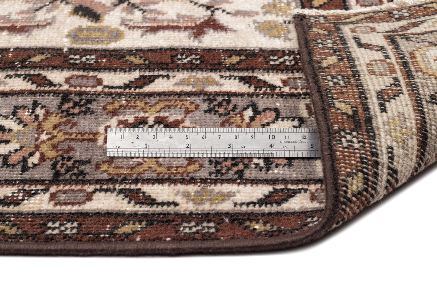 Traditional brown, cream, and grey patterned rug with a folded corner and a metal ruler for scale on a white background.