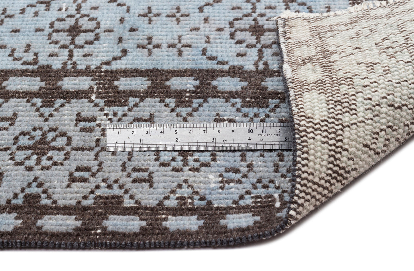 Light blue and dark brown rug with a geometric pattern, shown with a metal ruler for scale and a folded corner.