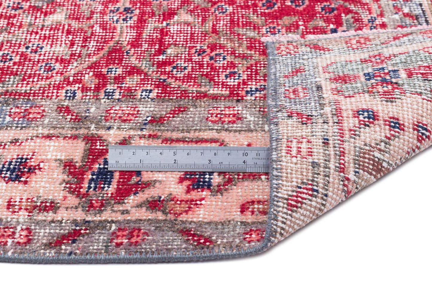 Red, pink, and blue distressed floral rug with a folded corner and a metal ruler for scale on a white background.