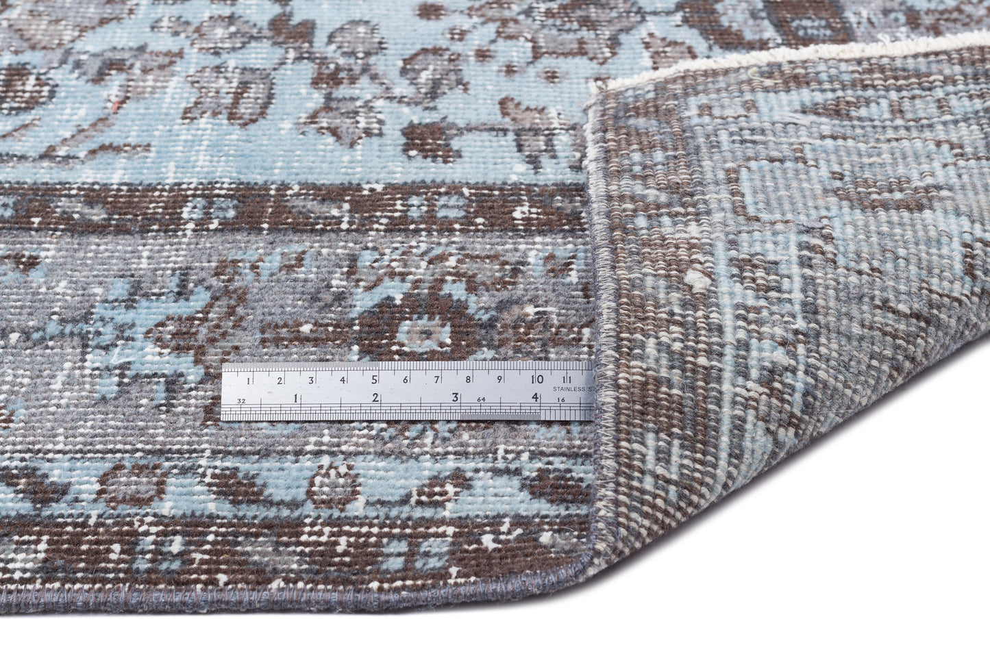Blue and brown distressed floral pattern rug with a folded corner and a metal ruler for scale on a white background.