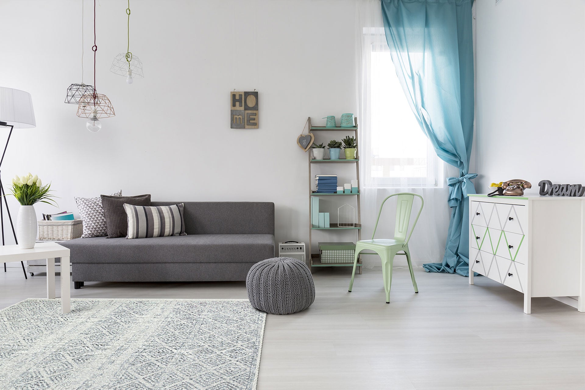 Modern living room featuring a gray sofa, Modern Mist - Washable Double-Sided Kilims - AR01 rug, white table, pouf, metal chair, geometric cabinet, shelves with plants, blue curtains, minimalist art on walls, and hanging lights.