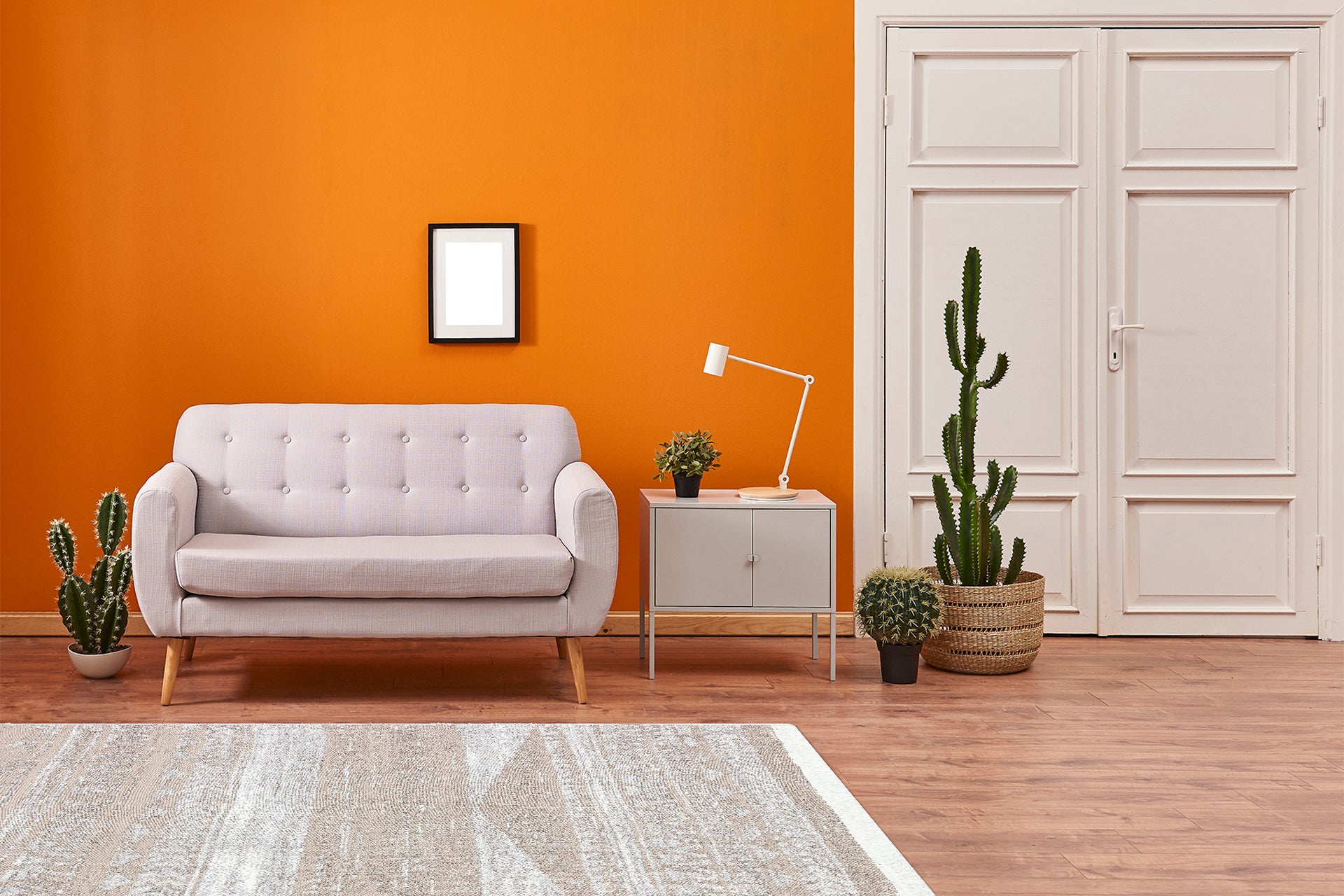 A modern living room with an orange wall, light gray sofa, side table and lamp, framed art, indoor cacti, white door, and the Earth Lines - Washable Double-Sided Kilims - AR06 allergy-friendly rug on a wooden floor.