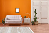 A modern living room with an orange wall, light gray sofa, side table and lamp, framed art, indoor cacti, white door, and the Earth Lines - Washable Double-Sided Kilims - AR06 allergy-friendly rug on a wooden floor.