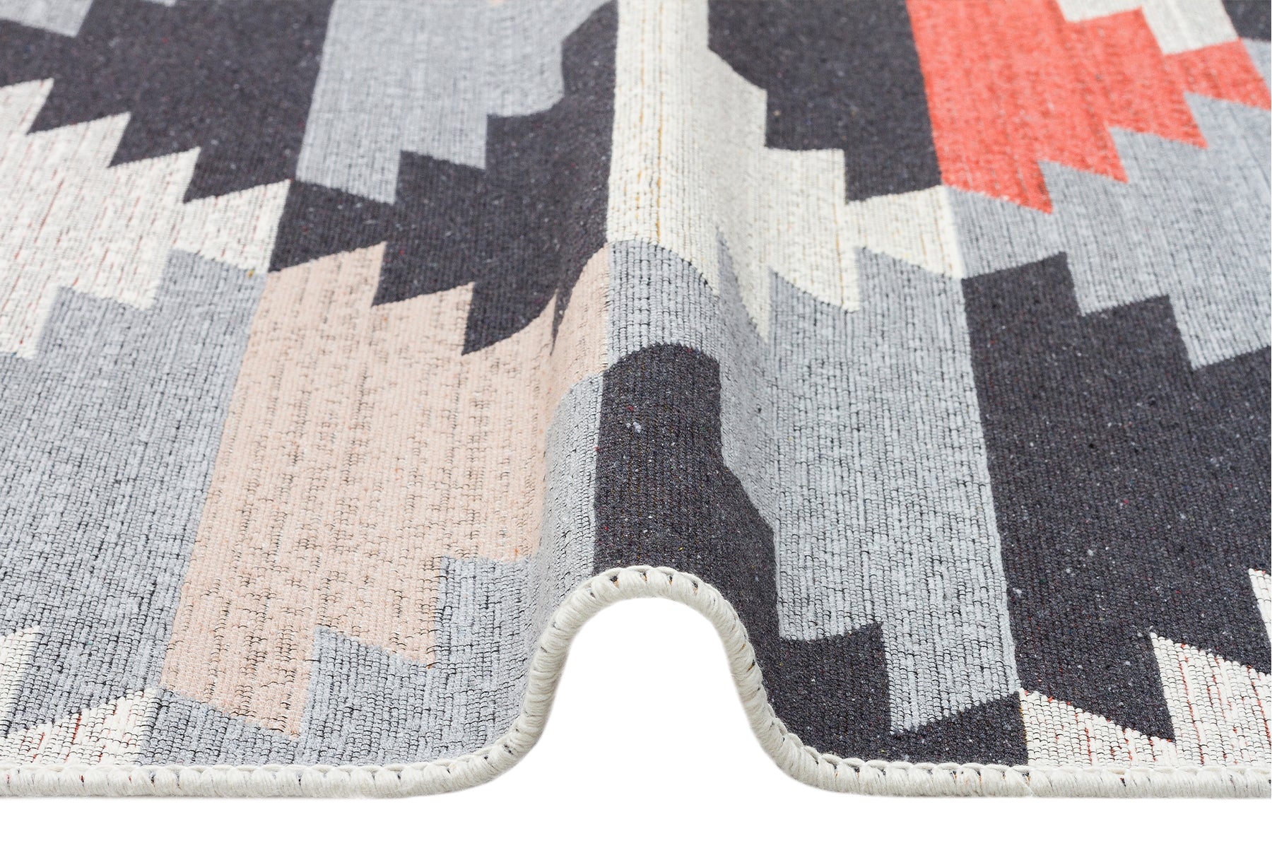 A close-up of the Artisan Touch - Washable Double-Sided Kilims - AR20 rug shows its geometric zigzag pattern in black, gray, beige, and coral tones with a wave-like fold and visible edge stitching.