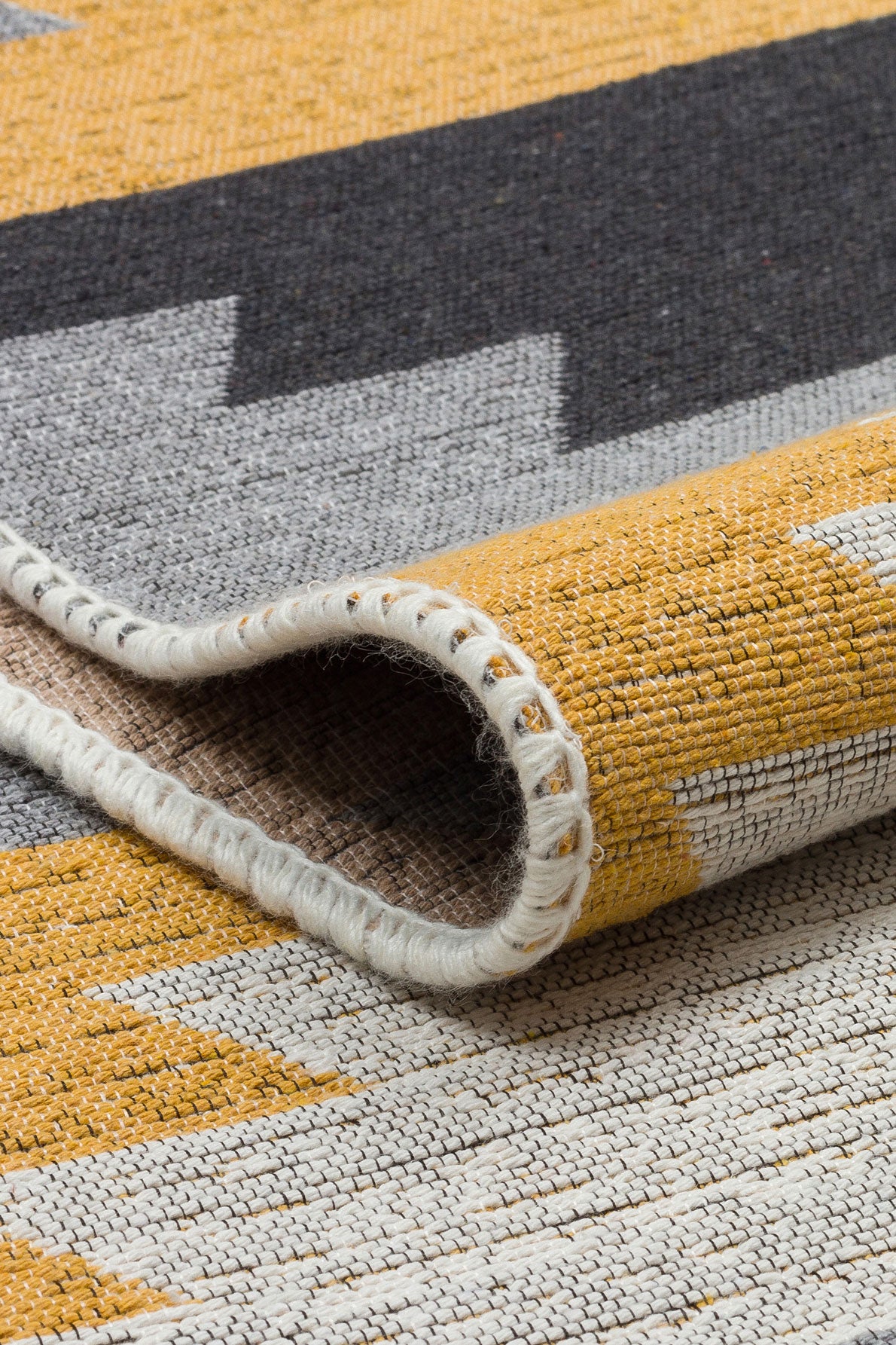 Close-up of the Artisan Touch - Washable Double-Sided Kilims - AR20 showing yellow, gray, black, and white geometric patterns. One lifted corner reveals its texture and detailed stitching along the edge.