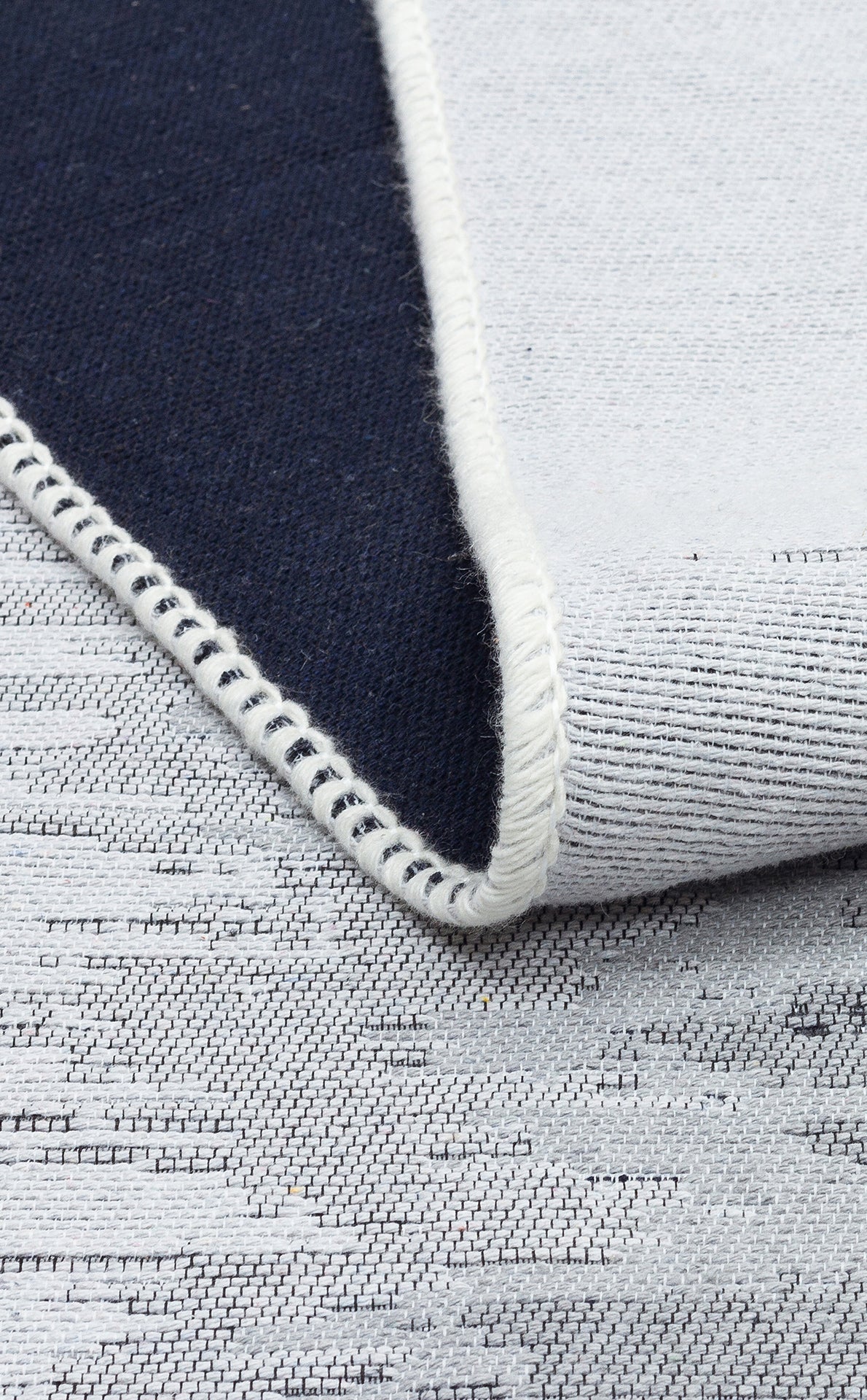 A close-up of the Charcoal Frame - Washable Double-Sided Kilims - AR40 shows a folded textile with one dark blue side, the other light gray with a subtle pattern, and an edge finished in white stitching.