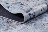 A close-up of the Abstract Atmosphere - Washable Rug - JR834 (Custom Size) in soft blue, gray, and beige tones. One corner is folded to show its dark textured underside—washable and ideal for high-traffic spaces.