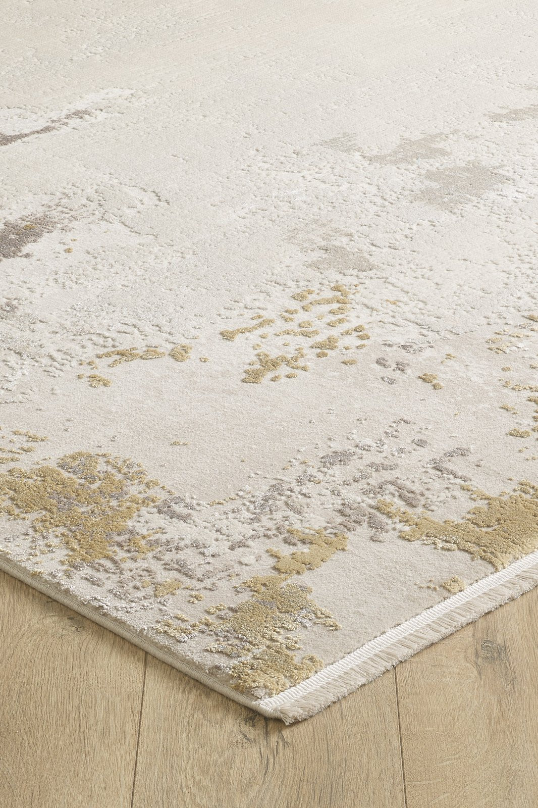 A close-up of the Abstract Blitz Premium Rug - Gold - M658F showcases its textured, abstract beige, tan, and light gray pattern on a light wood floor. The machine-made rug’s corner is slightly lifted, revealing its woven edge.