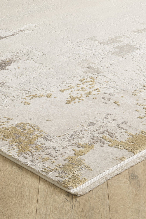 Load image 1 in gallery view. A close-up of the Abstract Blitz Premium Rug - Gold - M658F showcases its textured, abstract beige, tan, and light gray pattern on a light wood floor. The machine-made rug’s corner is slightly lifted, revealing its woven edge.