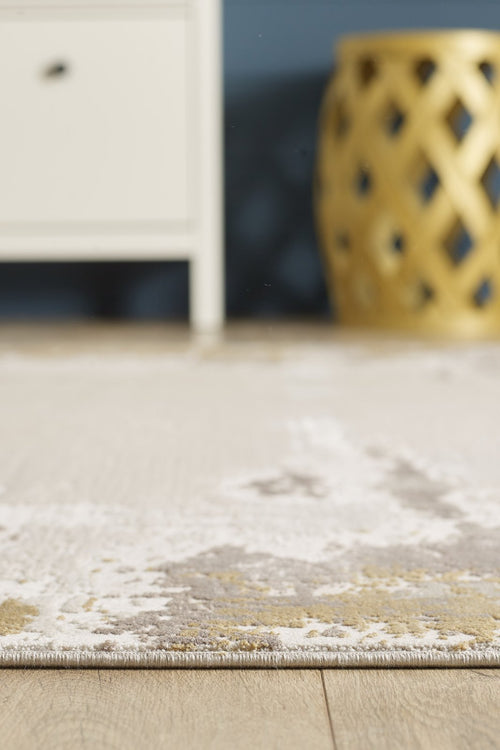 Load image 1 in gallery view. A close-up of the Abstract Blitz Premium Rug - Gold - M658F on a wooden floor, with part of a white cabinet on the left and a yellow, lattice-patterned cylindrical stool in the background.