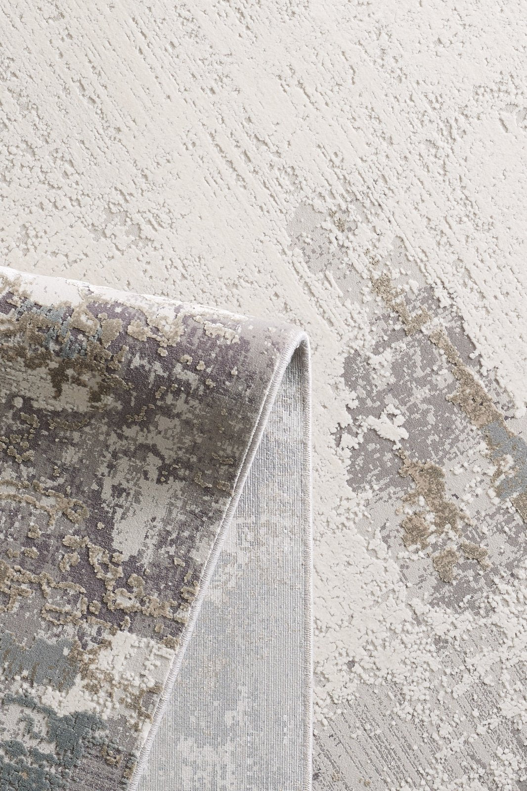 A close-up of the Abstract Blitz Premium Rug - Silver - M658D shows its modern abstract patterns in beige, gray, taupe, and muted blue, with one corner folded to reveal the machine-made weave and backing.