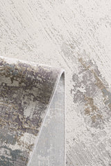 A close-up of the Abstract Blitz Premium Rug - Silver - M658D shows its modern abstract patterns in beige, gray, taupe, and muted blue, with one corner folded to reveal the machine-made weave and backing.