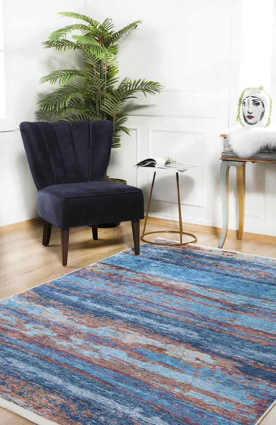 A modern living room with a blue accent chair, round white side table, plant, illustrated face pillow, and the Abstract Elegance - Machine-Washable Rug JR87 (Custom Size) on wood floors. Light pours in through a window.