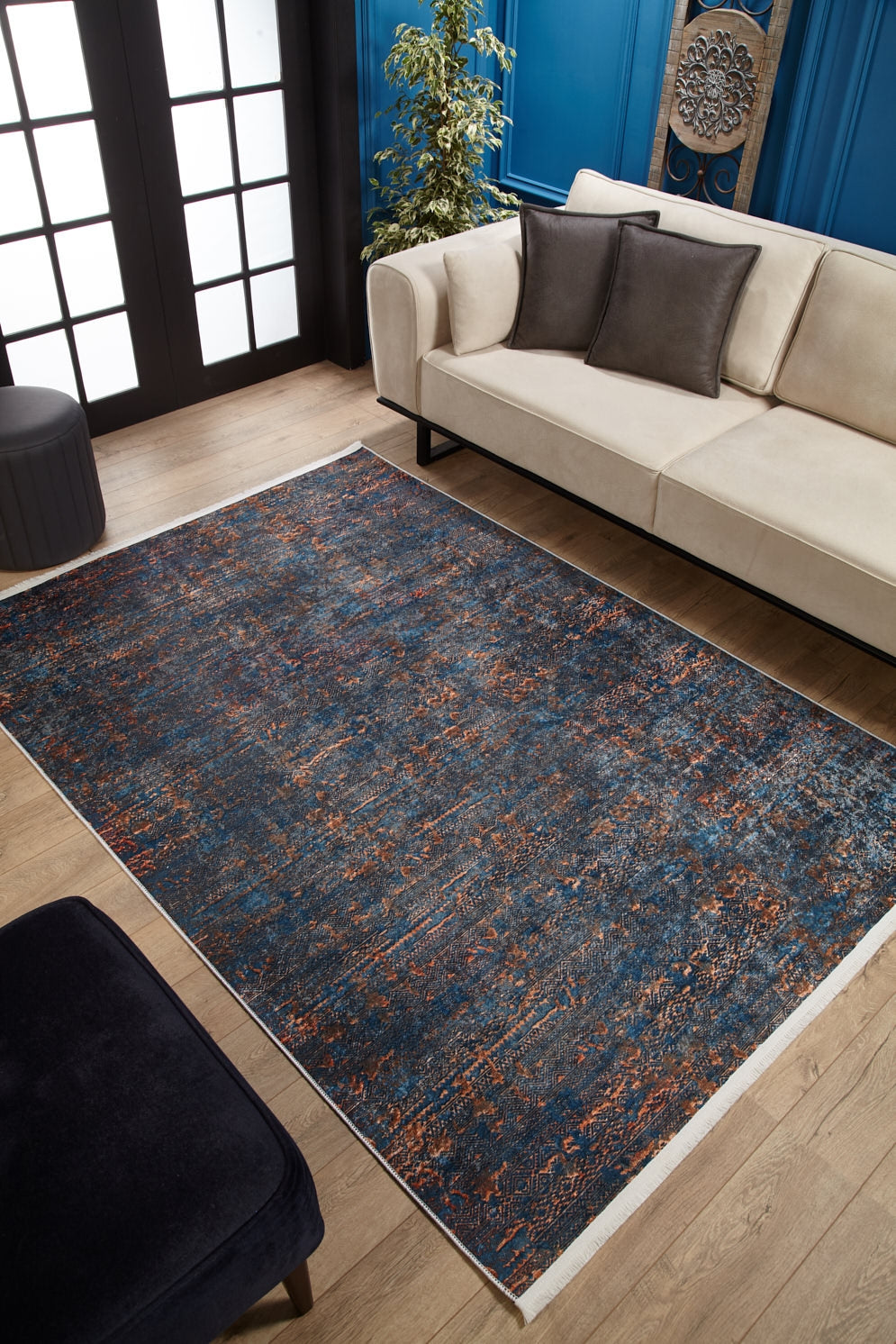 A modern living room features a beige sofa, dark cushions, wooden floor, a plant, blue walls with molding and art, and the Abstract Harmony - Machine-Washable Rug JR1882 (Custom Size) in blue and orange—ideal for high-traffic areas.