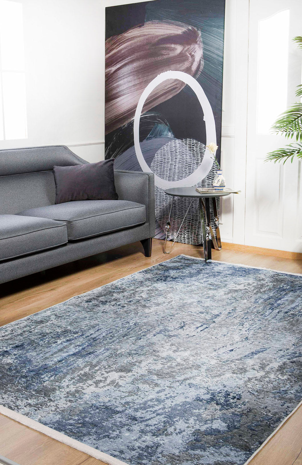 A modern living room with a gray sofa, the Abstract Oasis - Washable Rug - JR144 (Custom Size) in blue-gray, a side table with flowers, a large abstract painting, and a green potted plant in the corner.