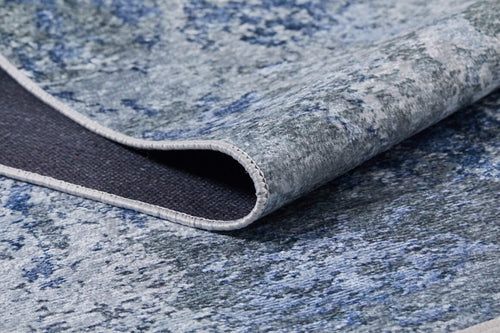 Load image 1 in gallery view. A close-up of the Abstract Oasis - Washable Rug - JR144 (Custom Size) in blue and gray shows its textured abstract pattern. One corner is lifted, revealing the dark backing and stitched edge of this allergy-friendly rug.