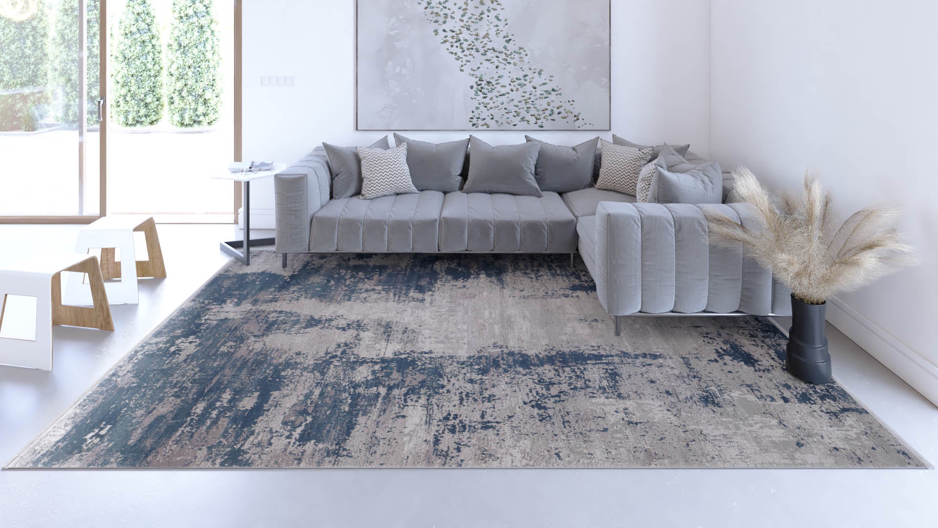 A modern living room features a gray sectional sofa with cushions, a side table, abstract wall art, the Abstracta Blue Premium Rug - 1004F in blue and beige, a black vase with pampas grass, and large windows overlooking greenery.