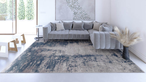 Load image 1 in gallery view. A modern living room features a gray sectional sofa with cushions, a side table, abstract wall art, the Abstracta Blue Premium Rug - 1004F in blue and beige, a black vase with pampas grass, and large windows overlooking greenery.