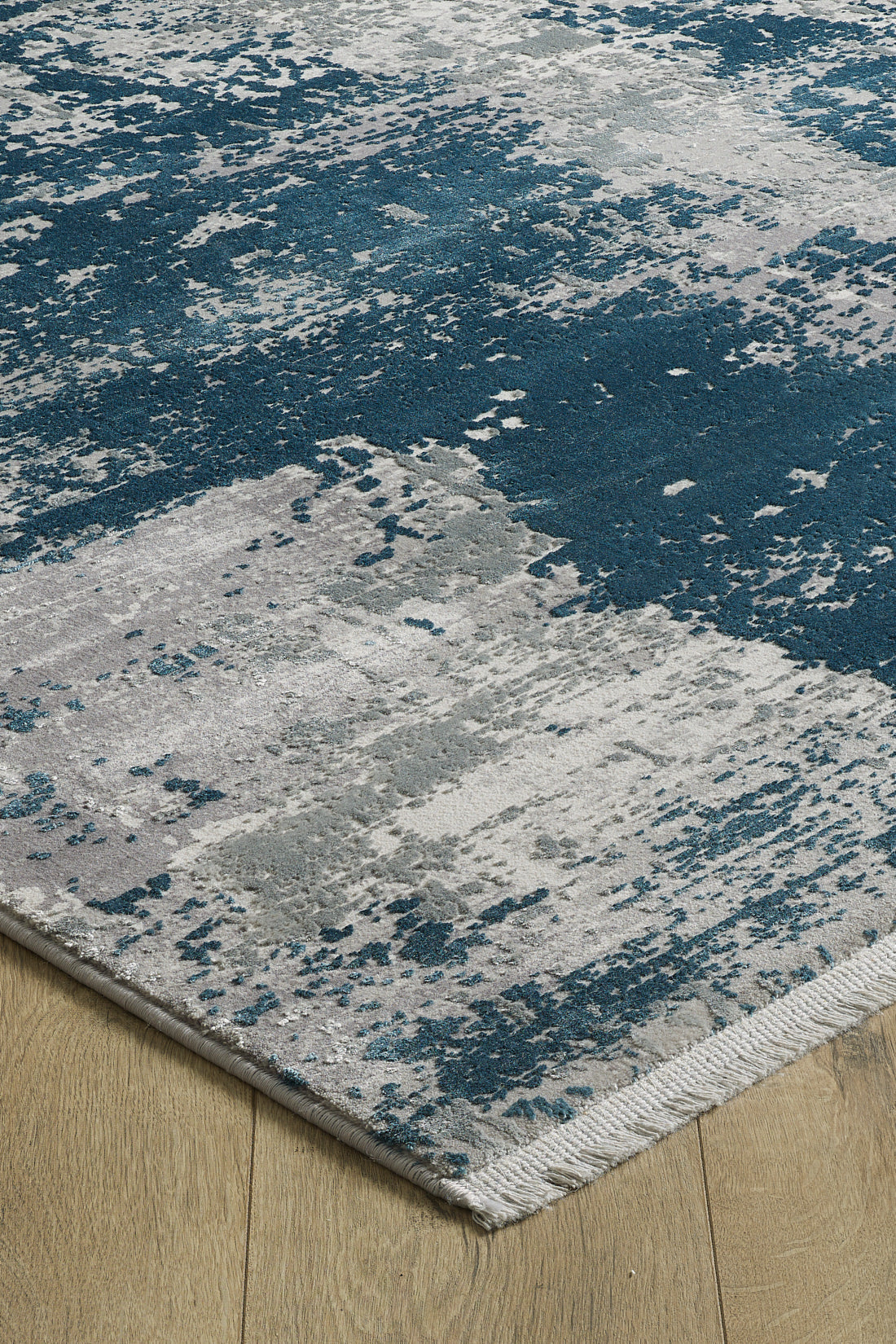 A close-up of the Abstracta Blue Premium Rug - 1004F shows its machine-made abstract pattern in blue, gray, and white on light wood flooring, highlighting the rug's corner and textured surface.