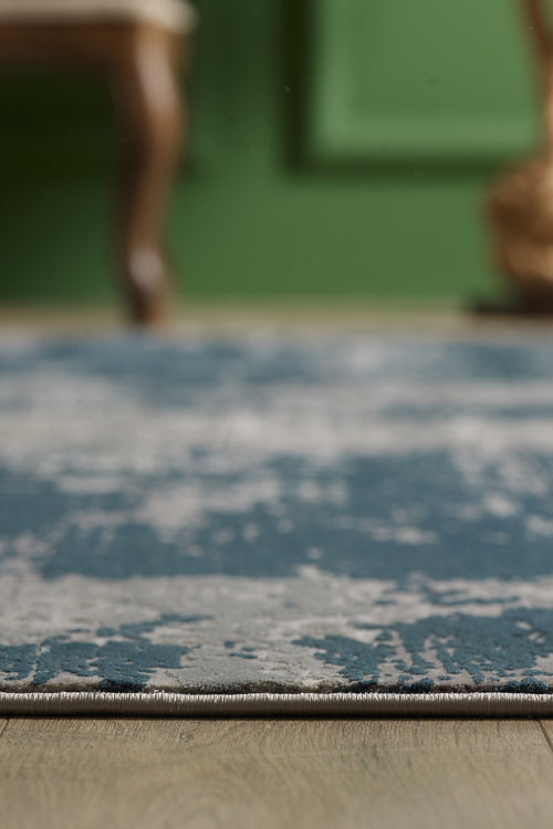 Load image 1 in gallery view. A close-up of the Abstracta Blue Premium Rug - 1004F, featuring blue and gray patterns on a wooden floor, with a blurred green wall and part of a chair in the background.