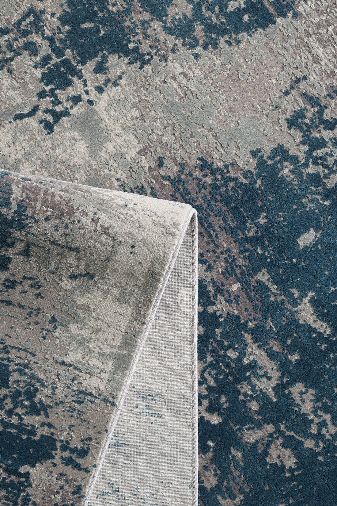 A close-up of the Abstracta Blue Premium Rug - 1004F, a machine-made piece with a folded corner, showcasing an abstract pattern in blue, gray, and beige tones.