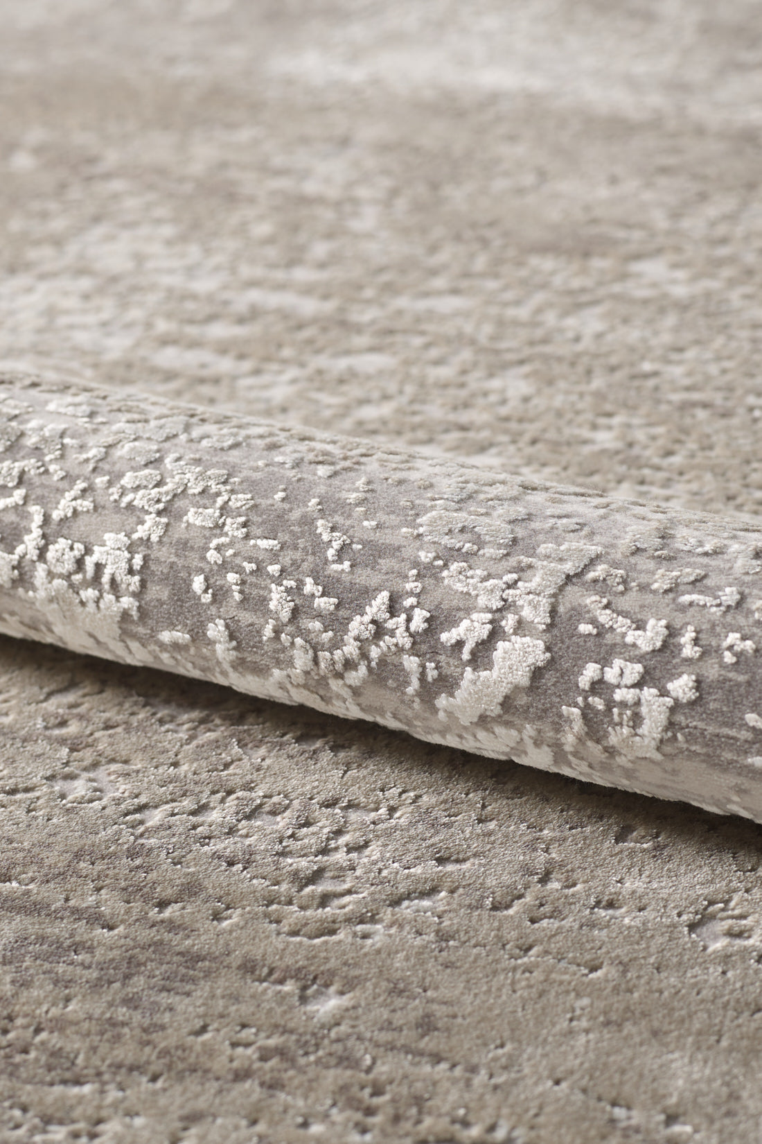 A close-up of the Abstracta Creme Premium Rug - 1004B, a rolled beige and light gray acrylic-viscose piece with a subtle abstract pattern on a matching carpeted surface.