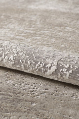 A close-up of the Abstracta Creme Premium Rug - 1004B, a rolled beige and light gray acrylic-viscose piece with a subtle abstract pattern on a matching carpeted surface.