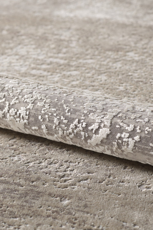 Load image 1 in gallery view. A close-up of the Abstracta Creme Premium Rug - 1004B, a rolled beige and light gray acrylic-viscose piece with a subtle abstract pattern on a matching carpeted surface.