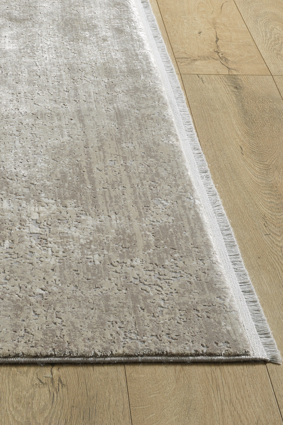 A close-up of the Abstracta Creme Premium Rug - 1004B shows its textured, light beige acrylic-viscose surface with subtle patterns and a fringed edge, placed on a light wooden floor. This is a machine-made rug.