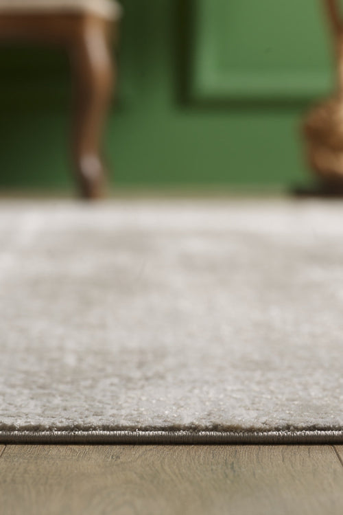 Load image 1 in gallery view. A close-up of the Abstracta Creme Premium Rug - 1004B, an acrylic-viscose piece in light gray on a wooden floor with a green wall and the blurred leg of a wooden chair in the background.