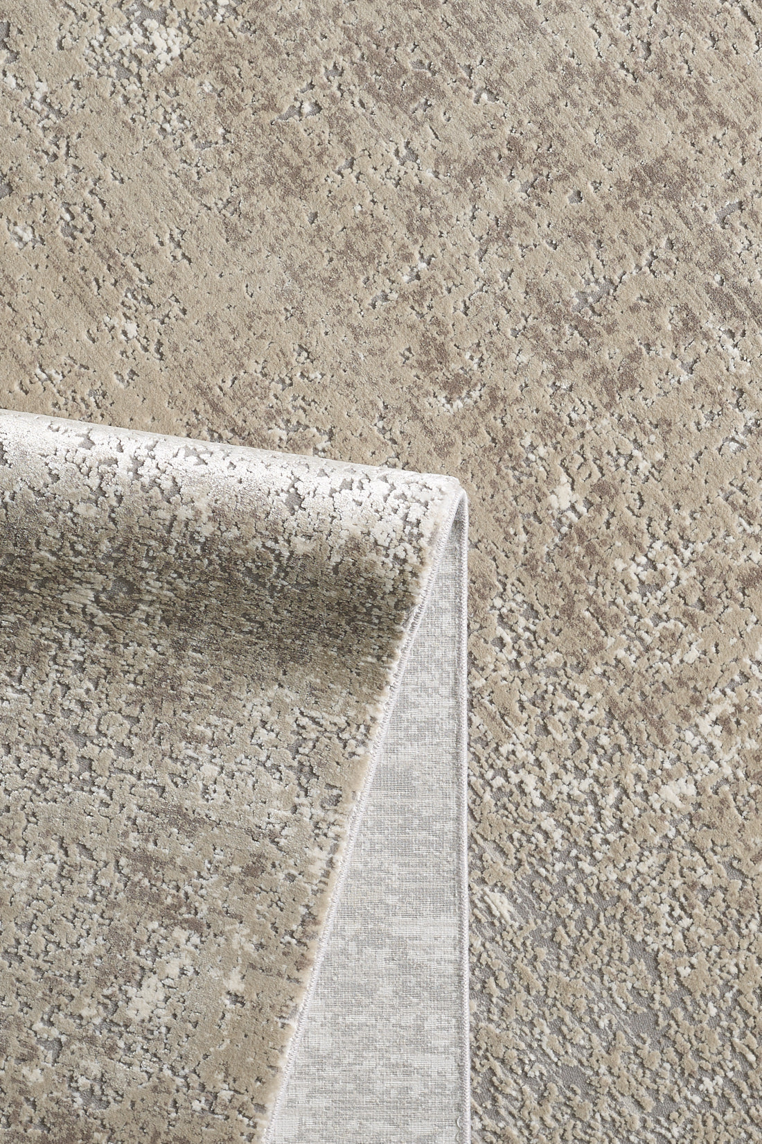 A close-up of the Abstracta Creme Premium Rug - 1004B showcases its beige and gray abstract speckled pattern, with a folded corner revealing the underside and edge stitching of this machine-made rug.