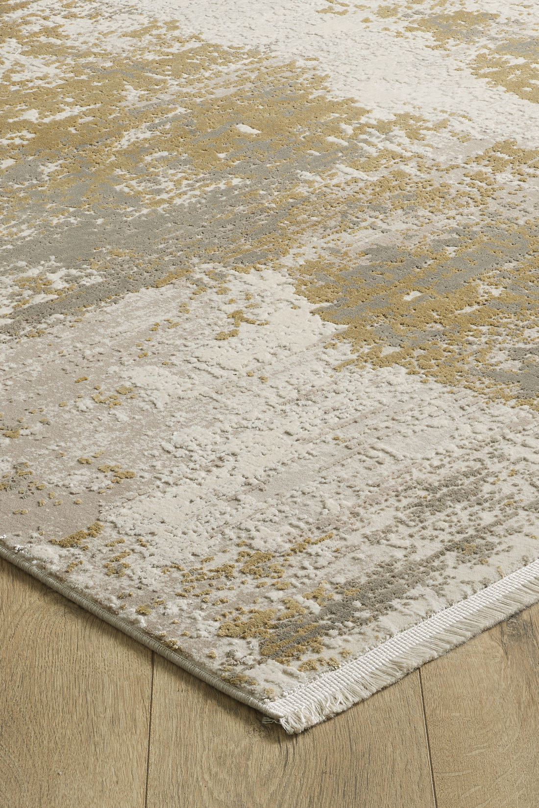 The Abstracta Gold Premium Rug - 1004G features a modern abstract pattern in beige, cream, gray, and gold tones with short fringe on one edge, shown here on a wooden floor.
