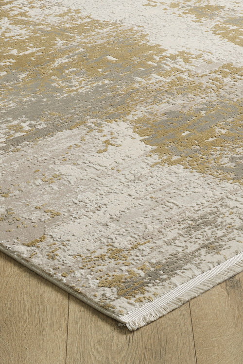 Load image 1 in gallery view. The Abstracta Gold Premium Rug - 1004G features a modern abstract pattern in beige, cream, gray, and gold tones with short fringe on one edge, shown here on a wooden floor.