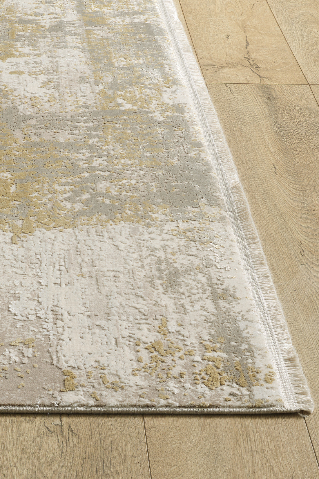 A close-up of the Abstracta Gold Premium Rug - 1004G, a rectangular machine-made rug with beige, grey, and yellow abstract patterns, textured surface, short fringes, and contemporary style on a light wood floor.