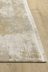 A close-up of the Abstracta Gold Premium Rug - 1004G, a rectangular machine-made rug with beige, grey, and yellow abstract patterns, textured surface, short fringes, and contemporary style on a light wood floor.