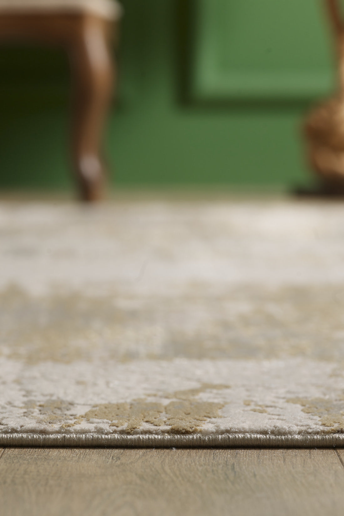 Close-up of the Abstracta Gold Premium Rug - 1004G in beige and light brown acrylic-viscose on a wooden floor, with a green wall and blurred wooden furniture in the background.