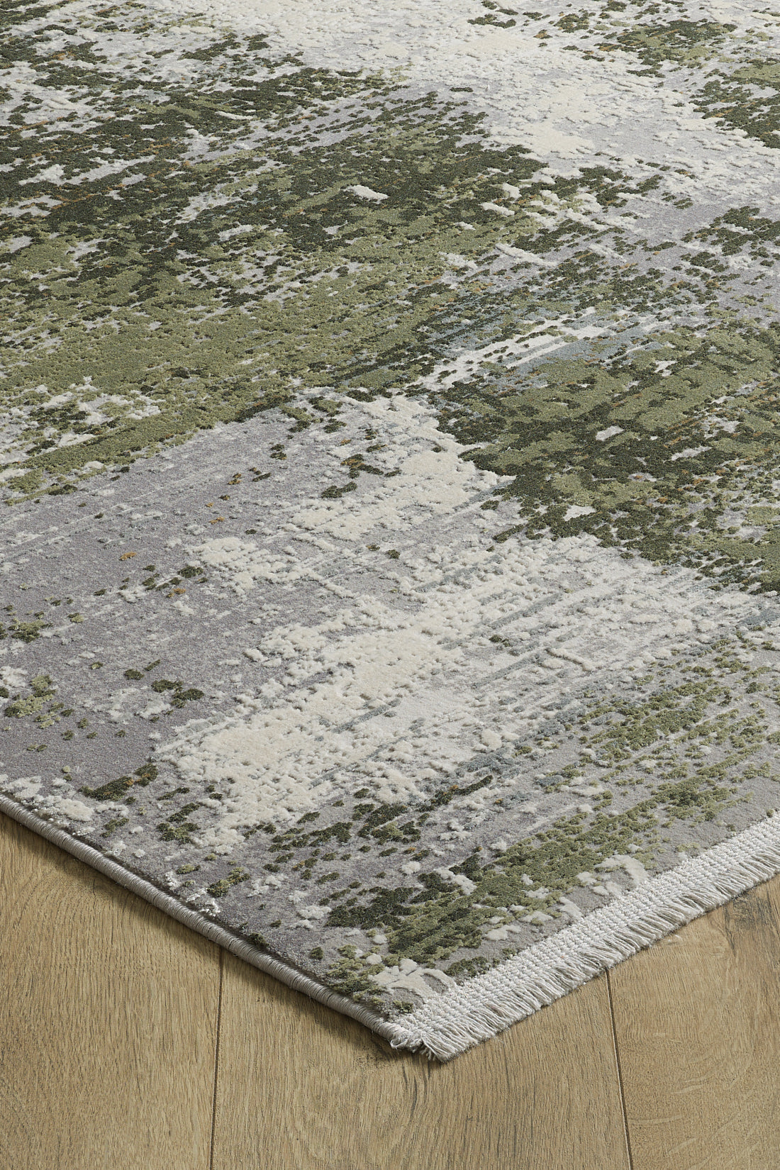 A close-up of the Abstracta Green Premium Rug - 1004H, a rectangular machine-made rug with an abstract green, gray, and white pattern, fringed edges, and a soft texture, placed on a light wooden floor for modern style.