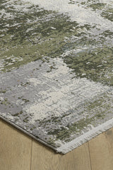 A close-up of the Abstracta Green Premium Rug - 1004H, a rectangular machine-made rug with an abstract green, gray, and white pattern, fringed edges, and a soft texture, placed on a light wooden floor for modern style.