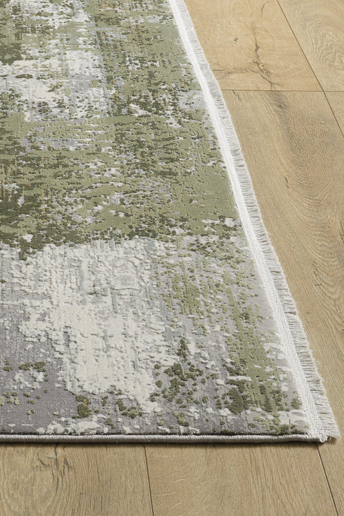 Load image 1 in gallery view. A close-up of the Abstracta Green Premium Rug - 1004H, a rectangular rug with green, white, and gray abstract patterns and white fringe along the edge, displayed on a light wooden floor.