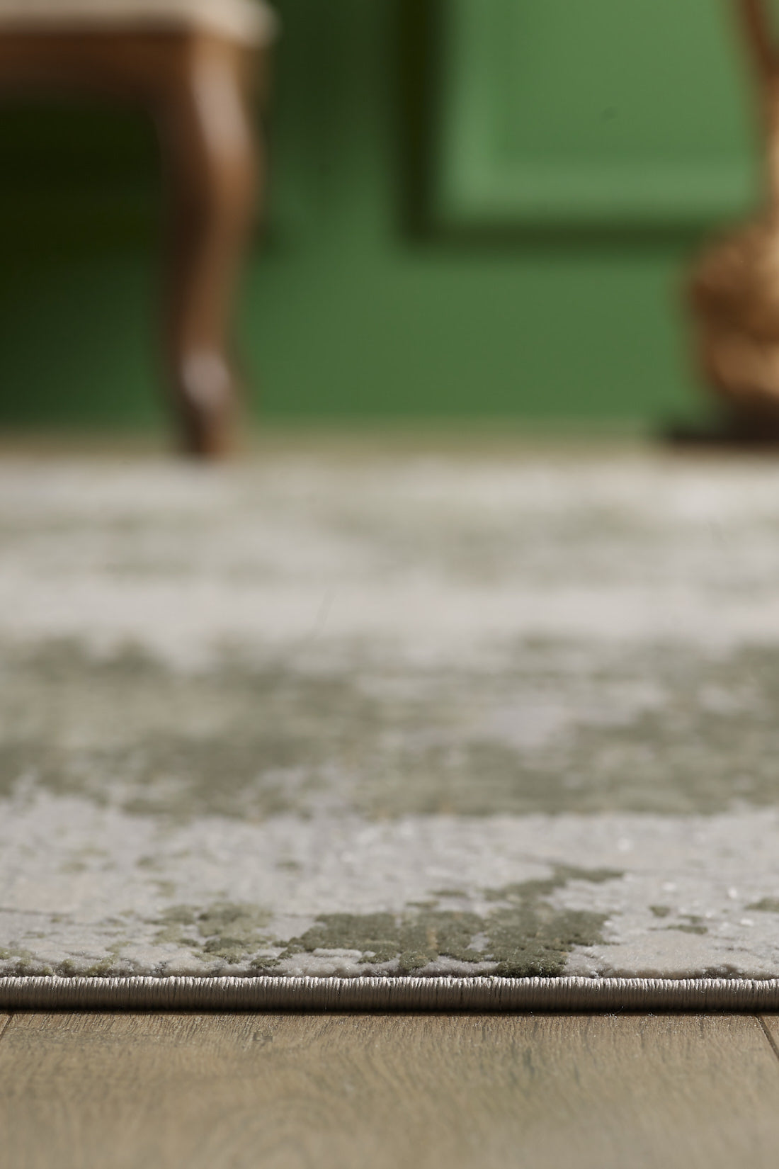 Close-up of the edge of the Abstracta Green Premium Rug - 1004H, a light-colored acrylic-viscose rug on a wooden floor, with a green wall and part of a wooden chair blurred in the background.