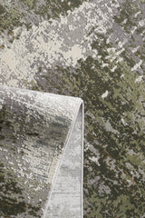 A close-up of the Abstracta Green Premium Rug - 1004H shows its abstract mottled green, gray, and cream pattern; one corner is folded to display the reverse side.