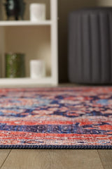 A close-up of the Aegis Handmade-Design Washable Rug - LCC3029 (Custom Size) with vibrant red, blue, and pink patterns on a wooden floor—perfect for allergy-sensitive homes. A white shelf and dark gray ottoman are in the background.