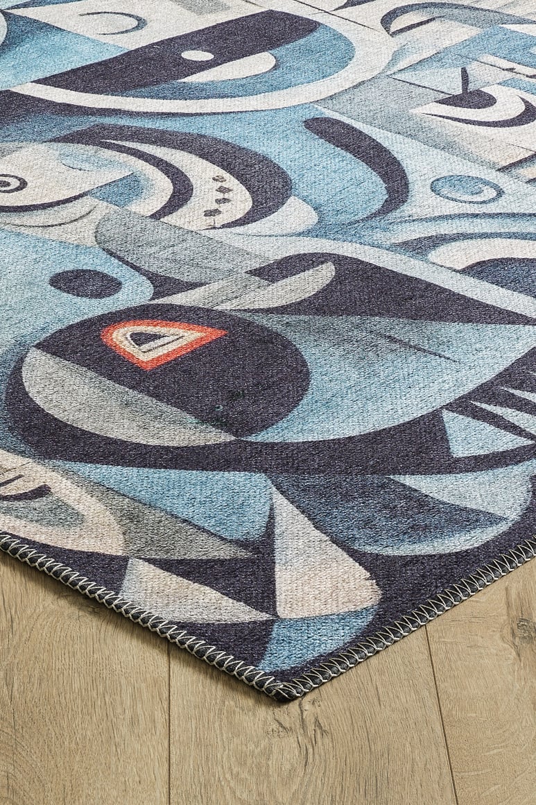 A close-up of the Aesthetic Anomaly Contemporary Washable Rug - MDJ004, showing blue tones with abstract white and black shapes plus a red and yellow triangle on light wood flooring. Features stitched edging—ideal for high-traffic areas.