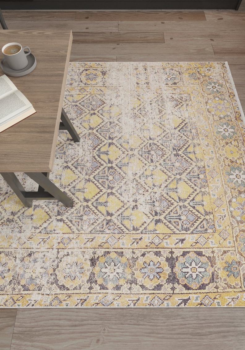 The Aged Splendor - Washable Rug - JR1906, a rectangular yellow-gold rug with faded gray geometric and floral design, is shown on a wooden floor by a light brown table with an open book and coffee—ideal for high-traffic spaces.
