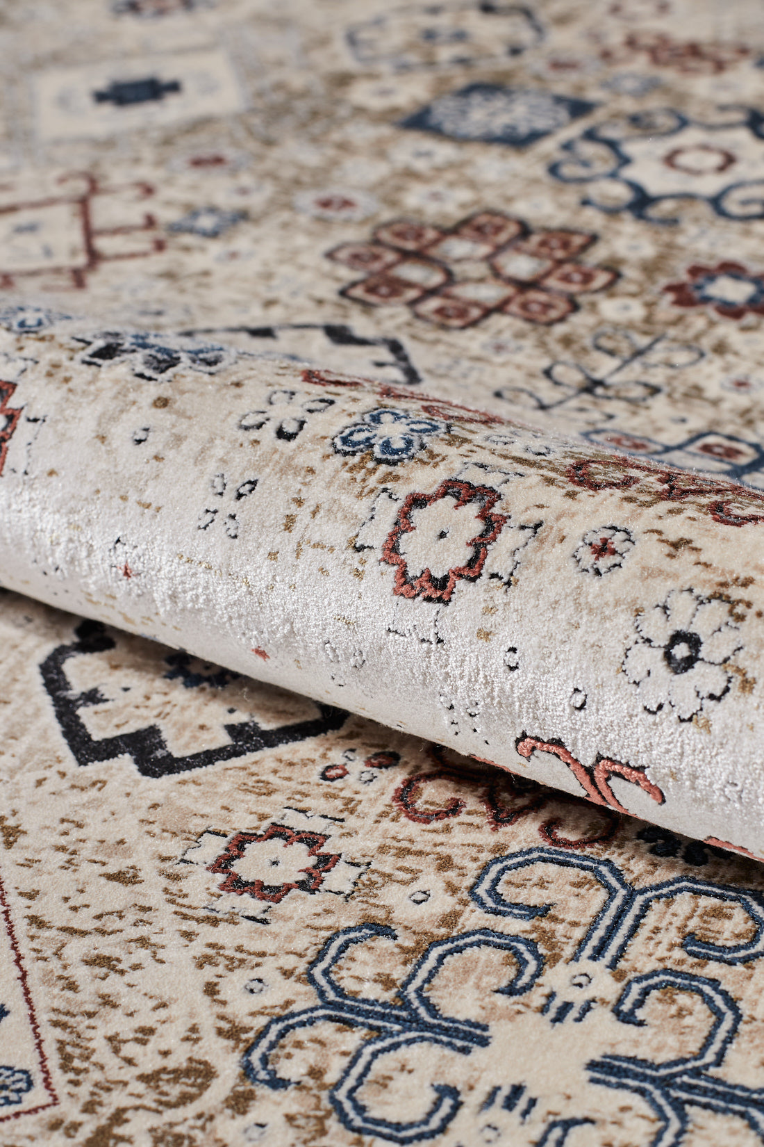 A close-up of the Aged Treasures Premium Rug - 2181A, a beige machine-made carpet featuring intricate multicolored geometric and floral patterns in blue, red, and brown. The rug is partially rolled to showcase its texture and detailed design.