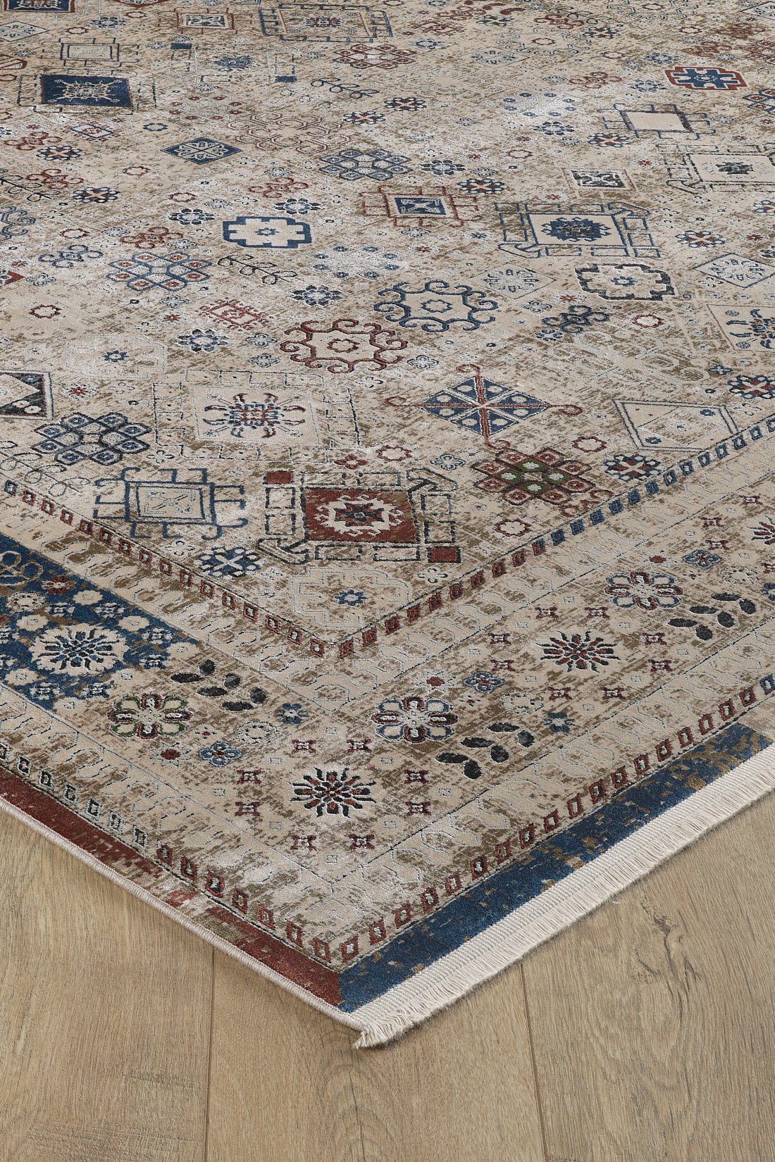 The Aged Treasures Premium Rug - 2181A is a beige geometric rug with blue, red, and brown patterns, a detailed border, and cream fringe. This machine-made rug lies on a light wooden floor for an elegant look.