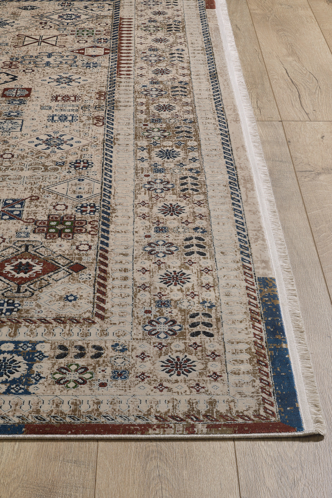 A close-up of the Aged Treasures Premium Rug - 2181A, a machine-made rug with intricate geometric and floral patterns in beige, blue, red, and brown on a light wood floor. Decorative borders accent its edges.